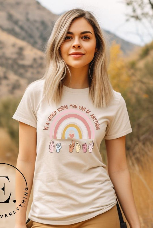 American sign language shirt with a rainbow and the phrase "In a world where you can be anything" and hands signing 'Be Kind' at the bottom on the rainbow on a tan colored shirt.