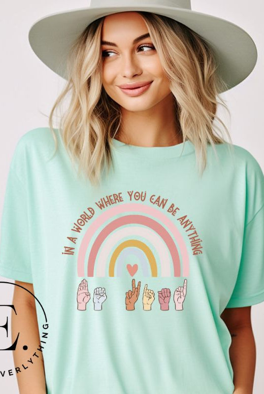 American sign language shirt with a rainbow and the phrase "In a world where you can be anything" and hands signing 'Be Kind' at the bottom on the rainbow on a teal colored shirt.