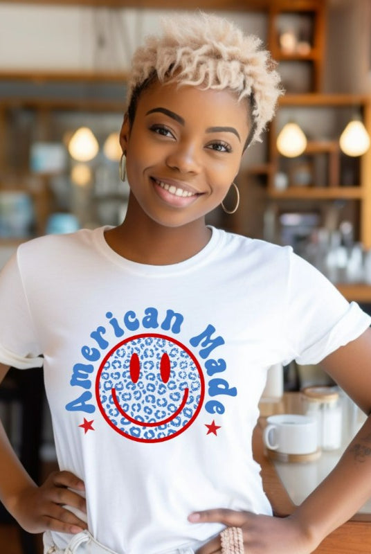 Close-up image of a USA July 4th graphic tee featuring the words 'American Made' surrounded by retro lettering around a bold blue cheetah print retro smiley face on the front. A playful and unique design perfect for celebrating July 4th in style on a white graphic tee. 