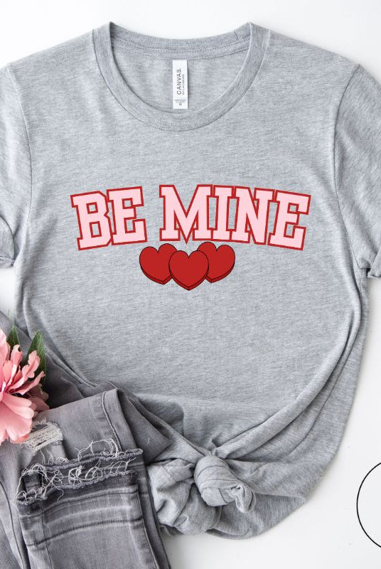 Elevate your Valentine's Day style with our "Be Mine" shirt! Featuring bold athletic lettering and three adorable hearts, on a grey shirt. 