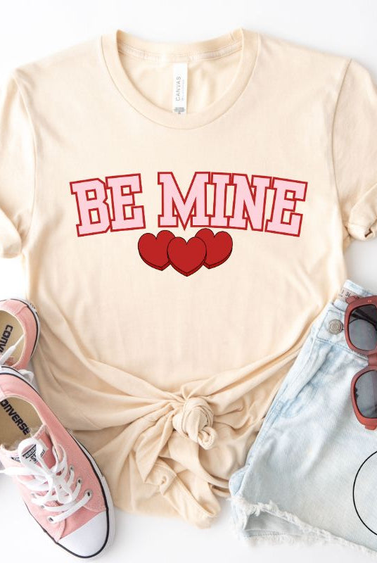 Elevate your Valentine's Day style with our "Be Mine" shirt! Featuring bold athletic lettering and three adorable hearts, on a soft cream shirt. 