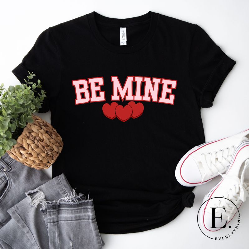 Elevate your Valentine's Day style with our "Be Mine" shirt! Featuring bold athletic lettering and three adorable hearts, on a black shirt. 