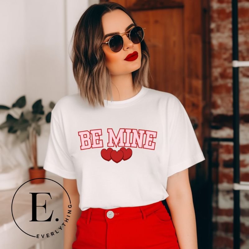 Elevate your Valentine's Day style with our "Be Mine" shirt! Featuring bold athletic lettering and three adorable hearts, on a white shirt. 
