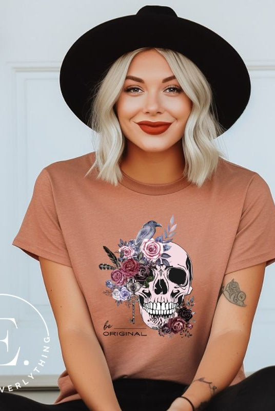 Looking for a unique Halloween shirt? Look no further! Our hauntingly beautiful shirt features a floral skull, raven, and the empowering slogan 'Be Original'. Stand out from the crowd with this unforgettable statement piece on a mauve shirt.