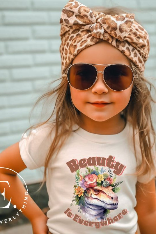 Unleash your child's wild side with our enchanting kids' shirt. Featuring a majestic dinosaur raptor adorned with a crown of flowers, this tee celebrates the beauty that surrounds us. With the inspiring message 'Beauty is Everywhere,' on a heather dust colored shirt. 
