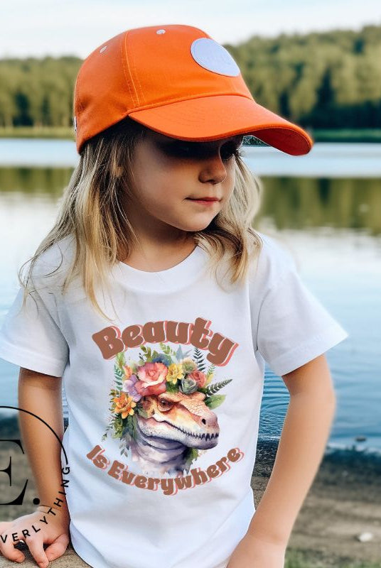 Unleash your child's wild side with our enchanting kids' shirt. Featuring a majestic dinosaur raptor adorned with a crown of flowers, this tee celebrates the beauty that surrounds us. With the inspiring message 'Beauty is Everywhere,' on a white shirt. 