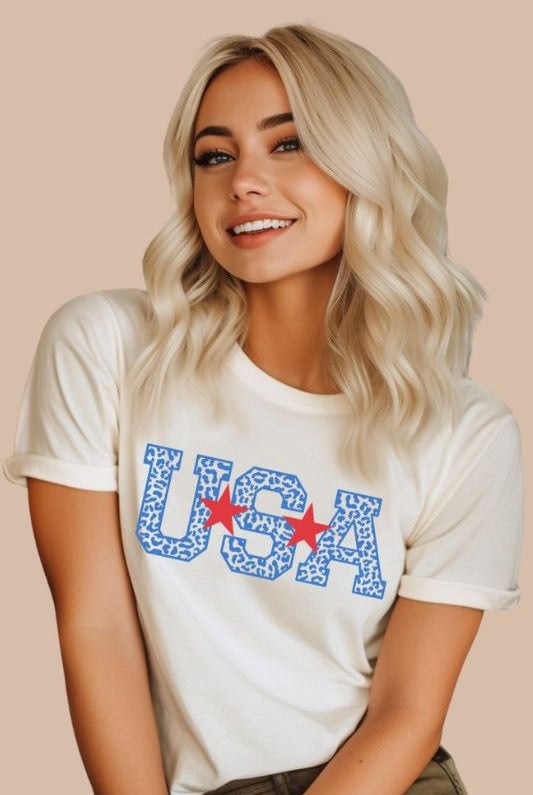 Stylish and bold USA July 4th graphic tee with blue cheetah print 'USA' on the front, adding a trendy and fierce touch to your patriotic wardrobe on a white graphic tee.