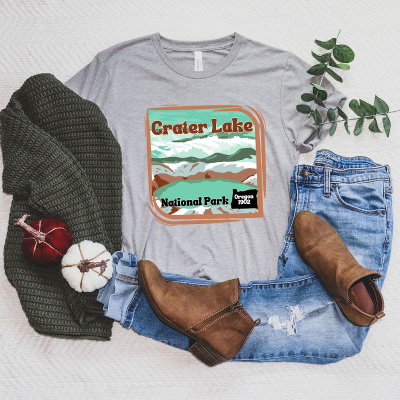 a colorful graphic of Crater Lake National Park, featuring mountains, water, and the text "Crater Lake National Park Oregon 1902 on a grey shirt. 