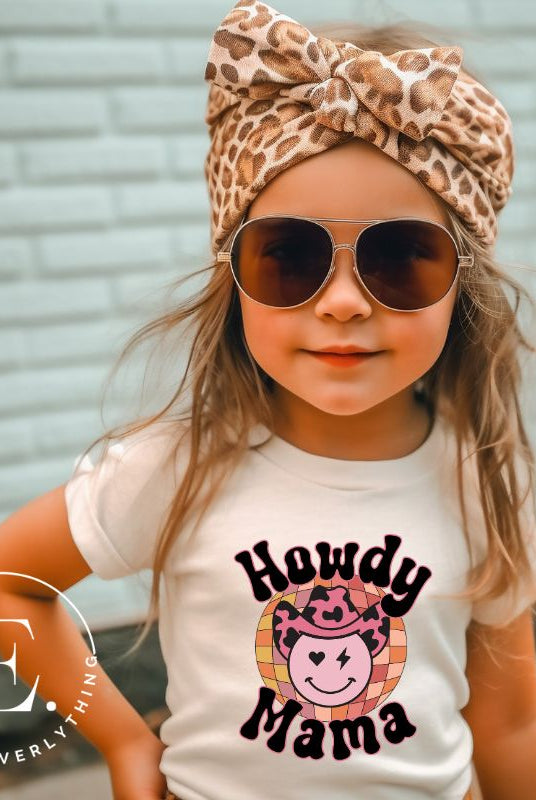 Yeehaw! Get ready to ride into style with our kids' shirt featuring a checkered ball with a winking smiley face, a lightning bolt, and a cowboy hat. With the playful saying 'Howdy Mama,' it's a fun and expressive choice on a heather dust colored shirt. 