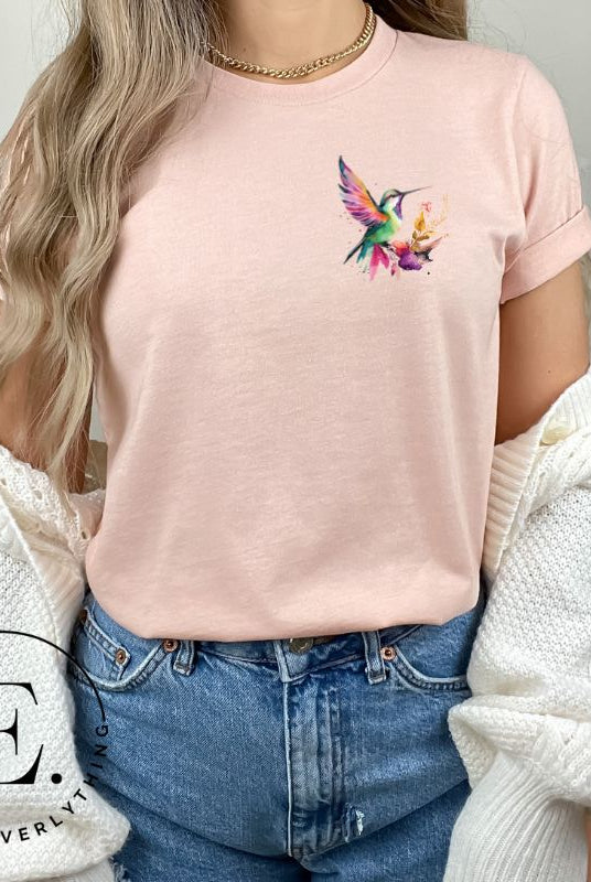 Elevate your style with our stunning t-shirt featuring a watercolor hummingbird delicately placed on the pocket on a peach shirt.