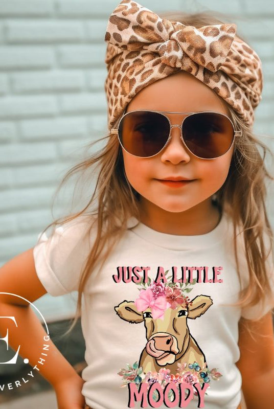 Our kid's shirt features an adorable highland cow with flowers and the quote 'Just a Little Moody,' adding humor and personality to the design on a white shirt. 