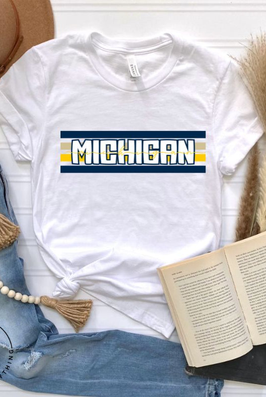 Elevate your collegiate style with our Michigan University graphic tee featuring iconic school colors and bold chest stripes. Emblazoned with "Michigan" in striking lettering, on a white shirt.