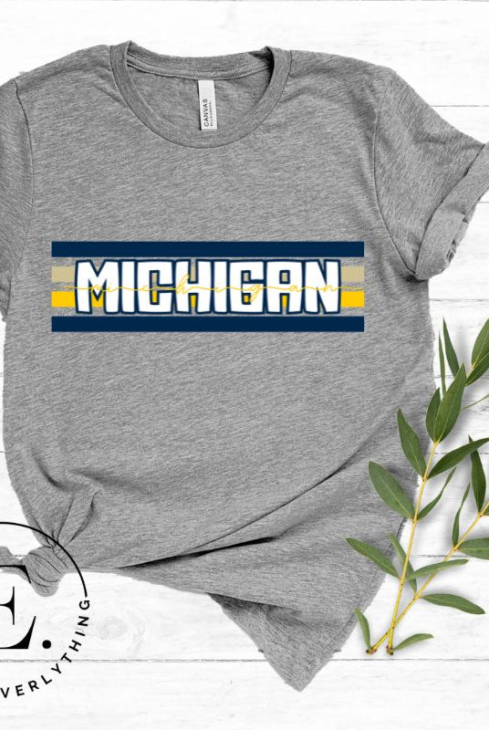 Elevate your collegiate style with our Michigan University graphic tee featuring iconic school colors and bold chest stripes. Emblazoned with "Michigan" in striking lettering, on a grey shirt. 