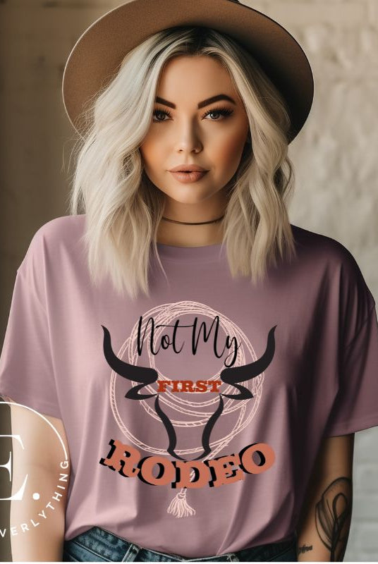 Unleash your cowboy spirit with our country western t-shirt boasting the statement "Not my First Rodeo" alongside bold bull horns and a lasso design on a mauve shirt. 