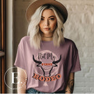 Unleash your cowboy spirit with our country western t-shirt boasting the statement "Not my First Rodeo" alongside bold bull horns and a lasso design on a mauve shirt. 