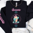 Black Gildan sweatshirt featuring a vibrant Cancer zodiac design. The front showcases an artistic representation of a woman with flowing hair adorned with a crab, surrounded by celestial elements like moons and stars, in pastel colors. The word "Cancer" is written in a stylish pink script above the design. The sleeve also has "Cancer" written vertically in matching pink script.