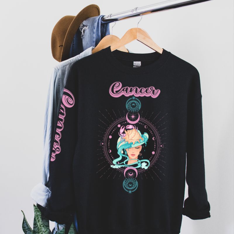 Black Gildan sweatshirt featuring a vibrant Cancer zodiac design. The front showcases an artistic representation of a woman with flowing hair adorned with a crab, surrounded by celestial elements like moons and stars, in pastel colors. The word "Cancer" is written in a stylish pink script above the design. The sleeve also has "Cancer" written vertically in matching pink script. The sweatshirt is displayed hanging on a clothing rack, accompanied by a denim jacket and a brown hat.