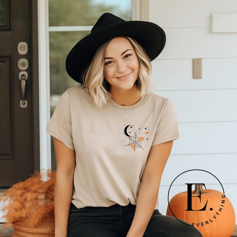 Embrace the enchanting night sky with our captivating t-shirt. Featuring a crescent moon, stars, and a spiderweb with three adorable spiders hanging down on the front pocket on a tan shirt. 