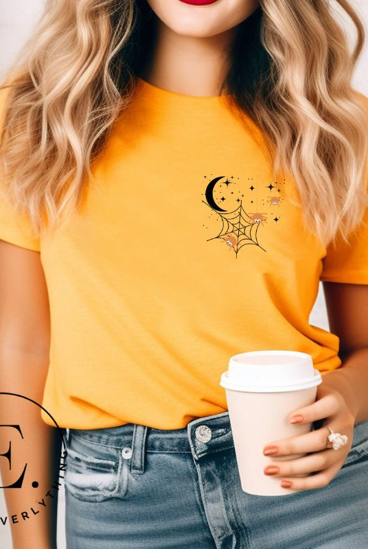 Embrace the enchanting night sky with our captivating t-shirt. Featuring a crescent moon, stars, and a spiderweb with three adorable spiders hanging down on the front pocket on an orange shirt. 