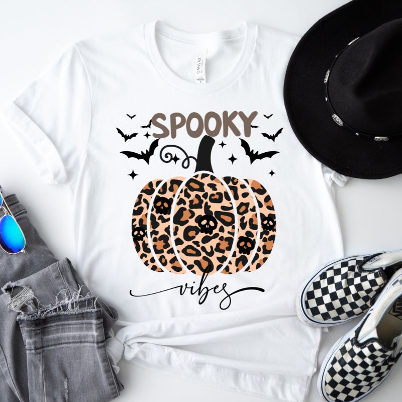 White t-shirt featuring a leopard-print pumpkin and "Spooky Vibes" text surrounded by bats, perfect for Halloween and fall season style.