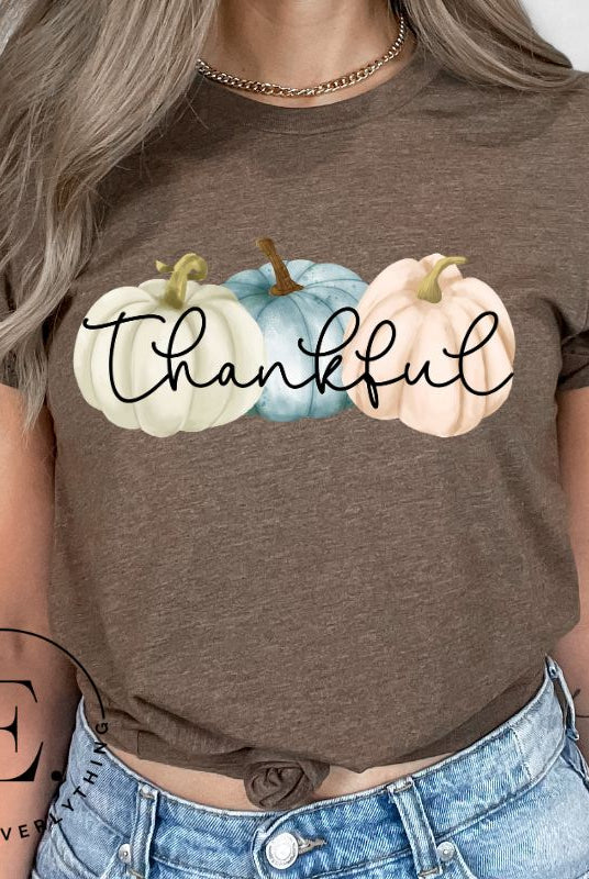 Express gratitude in style with our charming t-shirt. This design radiates autumn appreciation, featuring three pastel pumpkins and the word 'thankful' gracefully woven through the middle on a brown shirt. 