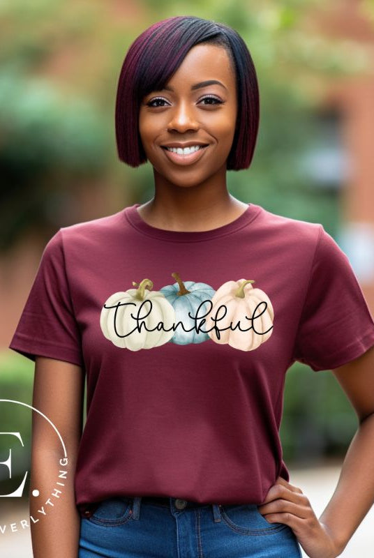 Express gratitude in style with our charming t-shirt. This design radiates autumn appreciation, featuring three pastel pumpkins and the word 'thankful' gracefully woven through the middle on a maroon shirt, 