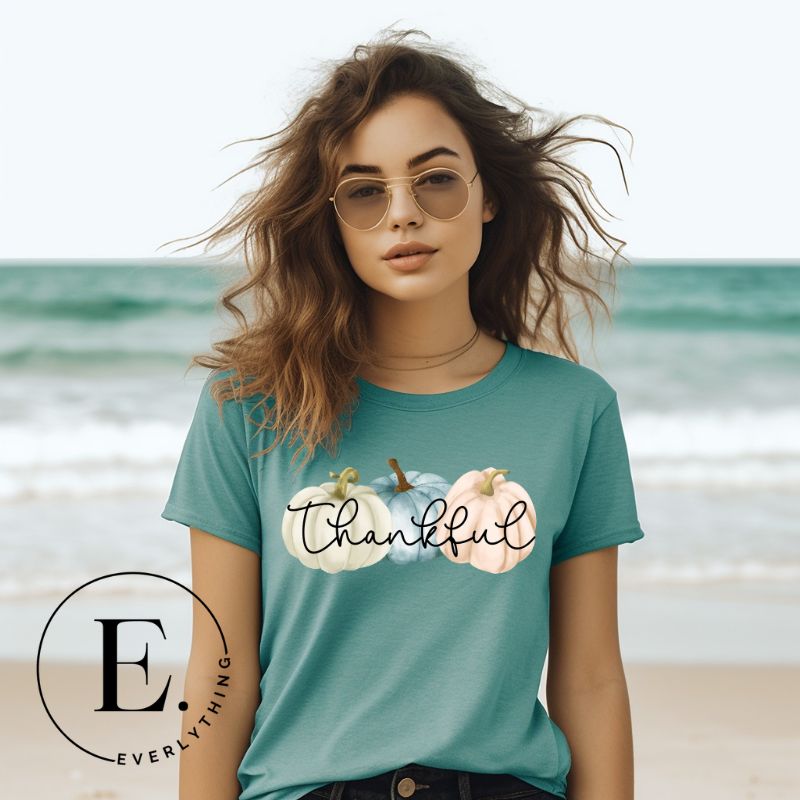 Express gratitude in style with our charming t-shirt. This design radiates autumn appreciation, featuring three pastel pumpkins and the word 'thankful' gracefully woven through the middle on a teal shirt.