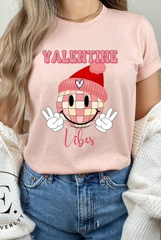 Get into the Valentine's Day spirit with our fun and funky shirt donning the words "Valentine Vibes" alongside a disco ball smiley face flashing peace fingers on a peach shirt. 