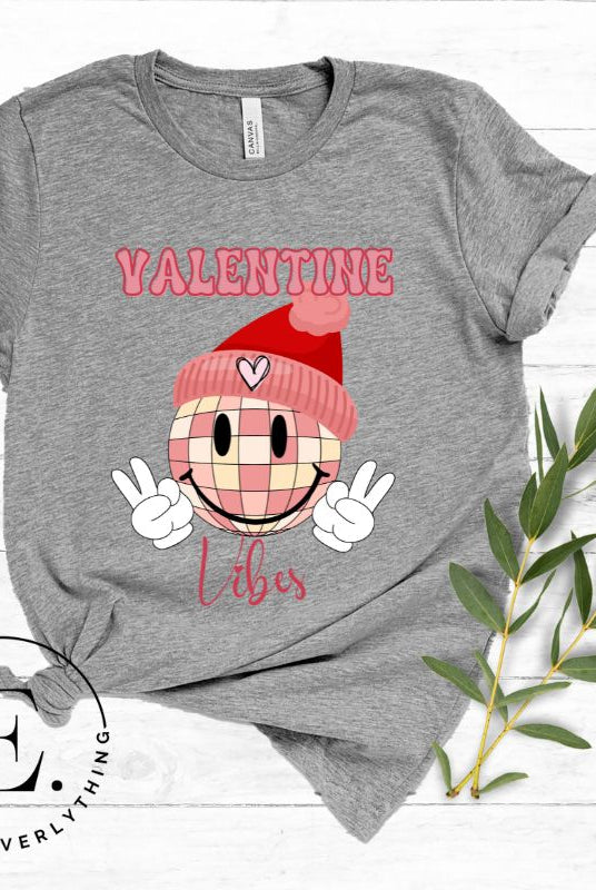 Get into the Valentine's Day spirit with our fun and funky shirt donning the words "Valentine Vibes" alongside a disco ball smiley face flashing peace fingers on a grey shirt. 