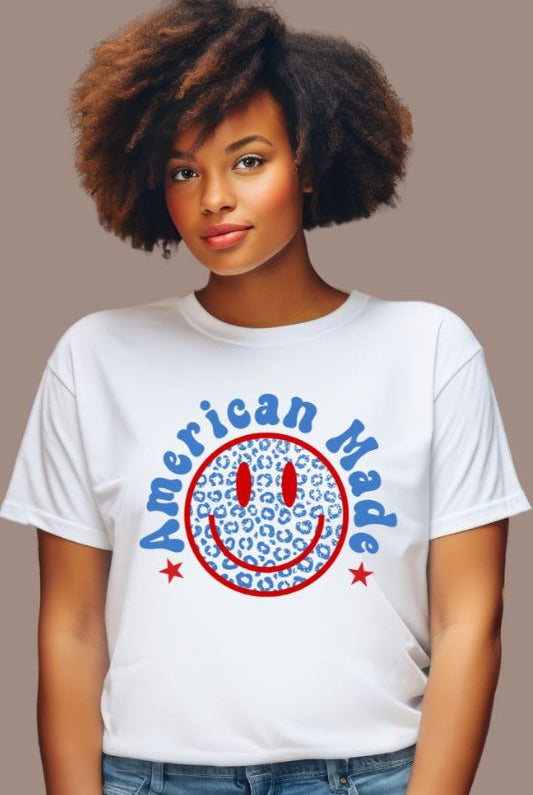 Close-up image of a USA July 4th graphic tee featuring the words 'American Made' surrounded by retro lettering around a bold blue cheetah print retro smiley face on the front. A playful and unique design perfect for celebrating July 4th in style on a white graphic tee.