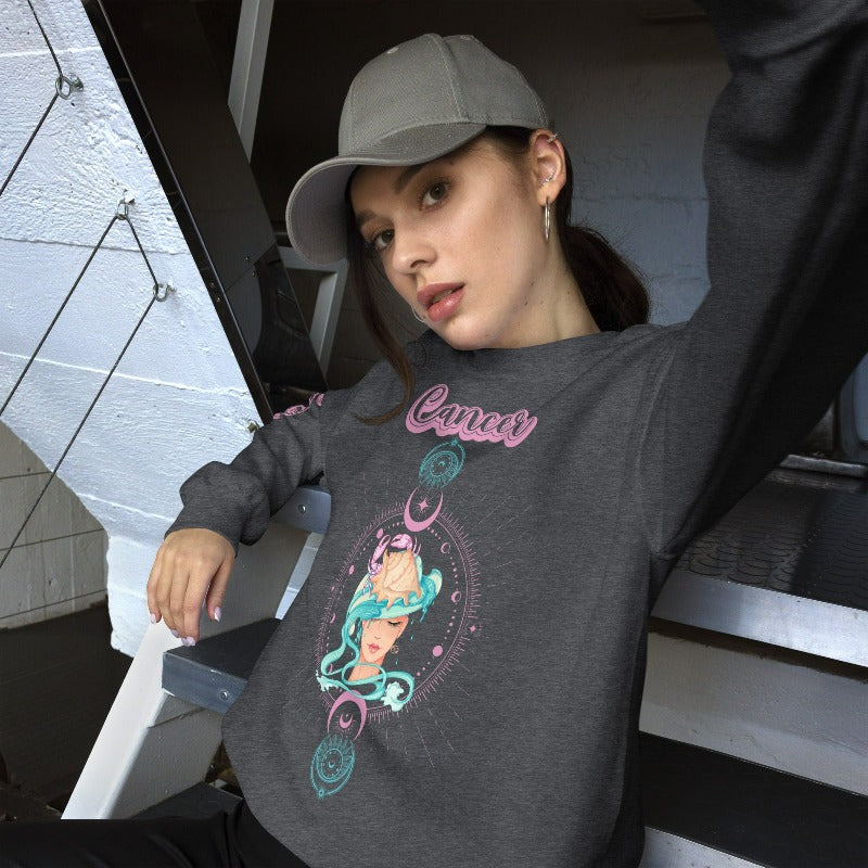 Dark Grey Gildan sweatshirt featuring a vibrant Cancer zodiac design. The front showcases an artistic representation of a woman with flowing hair adorned with a crab, surrounded by celestial elements like moons and stars, in pastel colors. The word "Cancer" is written in a stylish pink script above the design. The sleeve also has "Cancer" written vertically in matching pink script.