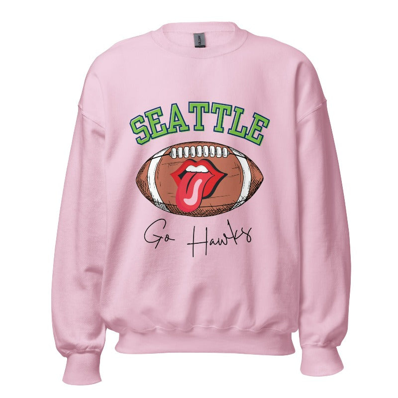 Support the Seattle Seahawks in style with this unique sweatshirt featuring a football and playful lips and tongue design. Featuring the team's slogan "Go Hawks" and the iconic Seattle wordmark, on a light pink sweatshirt. 