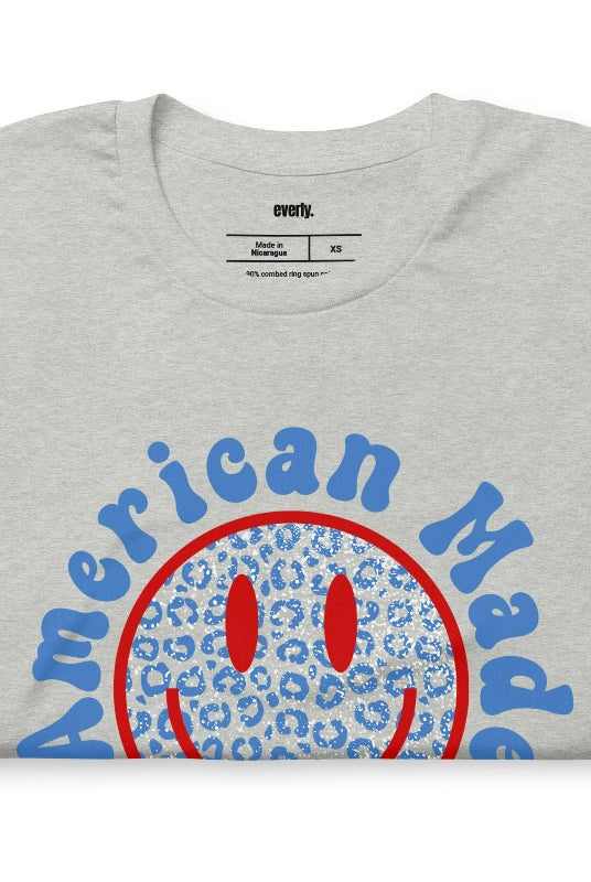 Close-up image of a USA July 4th graphic tee featuring the words 'American Made' surrounded by retro lettering around a bold blue cheetah print retro smiley face on the front. A playful and unique design perfect for celebrating July 4th in style on an athletic heather grey shirt. 