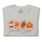  'Tis The Season' design with fall-themed graphics including a cupcake, maple leaf, football, and pumpkin, surrounded by falling leaves on a grey shirt. 