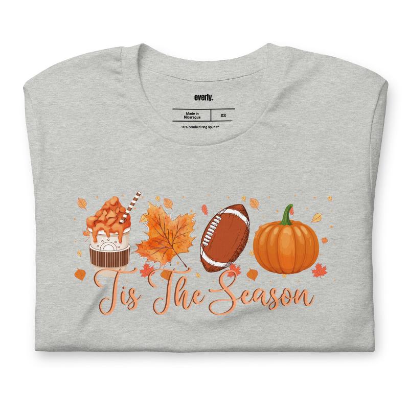  'Tis The Season' design with fall-themed graphics including a cupcake, maple leaf, football, and pumpkin, surrounded by falling leaves on a grey shirt. 