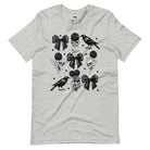 A grey unisex t-shirt featuring a Halloween-themed design with black roses, skulls, ravens, bows, and stars. The design is printed in black and white, giving it a gothic and spooky vibe.