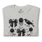 A grey unisex t-shirt featuring a Halloween-themed design with black roses, skulls, ravens, bows, and stars. The design is printed in black and white, giving it a gothic and spooky vibe.