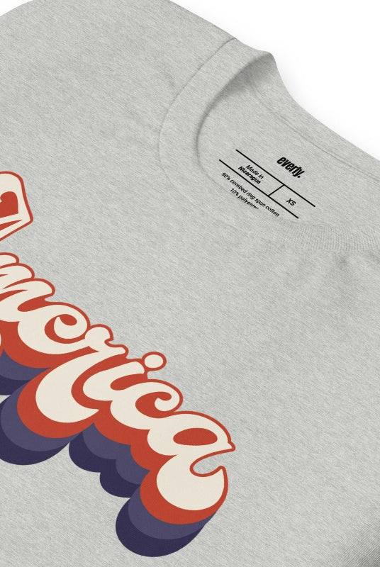 Close-up image of a USA July 4th graphic tee with the word 'America' spelled out in funky retro lettering on the front. This vintage-inspired tee is perfect for adding a fun and nostalgic touch to your Independence Day celebrations on an athletic heather grey graphic tee.