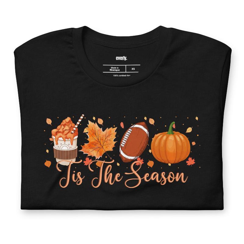  'Tis The Season' design with fall-themed graphics including a cupcake, maple leaf, football, and pumpkin, surrounded by falling leaves on a black shirt. 