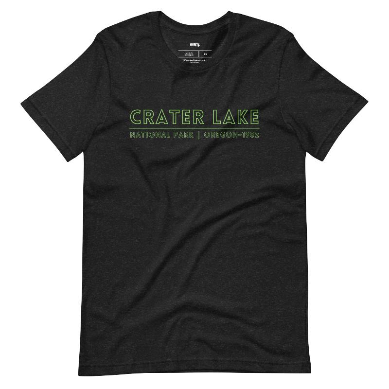 A vibrant design of Crater Lake National Park on the back, showcasing the lake and surrounding mountains in colorful hues, with the text "Crater Lake National Park | Oregon 1902" on the front. This design is on a black shirt. 