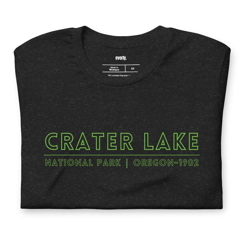 A vibrant design of Crater Lake National Park on the back, showcasing the lake and surrounding mountains in colorful hues, with the text "Crater Lake National Park | Oregon 1902" on the front. This design is on a black shirt. 