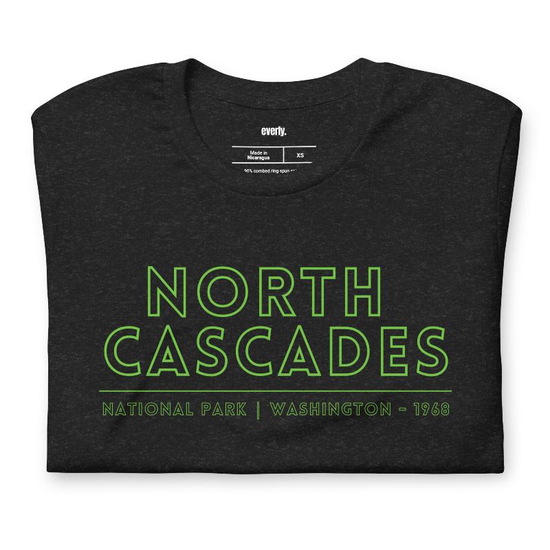 a vibrant, retro-style design of North Cascades National Park on the back, and "North Cascades National Park | Washington - 1968" text on the front on a black shirt.