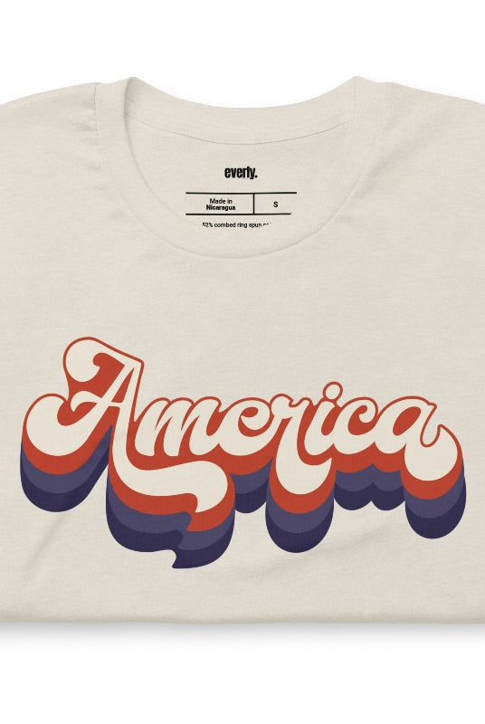 Close-up image of a USA July 4th graphic tee with the word 'America' spelled out in funky retro lettering on the front. This vintage-inspired tee is perfect for adding a fun and nostalgic touch to your Independence Day celebrations on a heather dust graphic tee. 