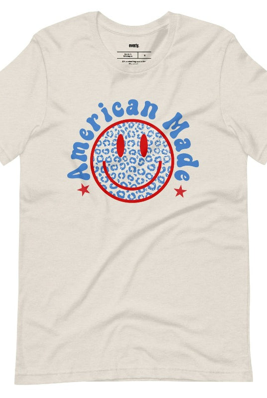 Close-up image of a USA July 4th graphic tee featuring the words 'American Made' surrounded by retro lettering around a bold blue cheetah print retro smiley face on the front. A playful and unique design perfect for celebrating July 4th in style on a heather dust graphic tee.