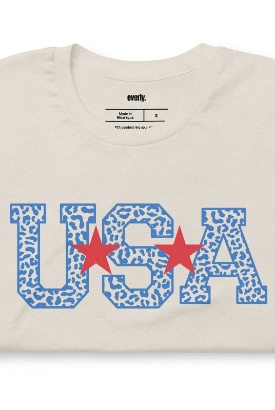 Stylish and bold USA July 4th graphic tee with blue cheetah print 'USA' on the front, adding a trendy and fierce touch to your patriotic wardrobe on a heather dust graphic tee.