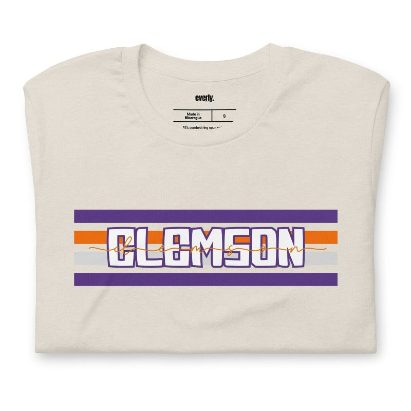 Bella + Canvas 3001 unisex shirt with Clemson University stripe design in purple, orange, and white, with "Clemson" written in bold letters on a beige graphic tee.