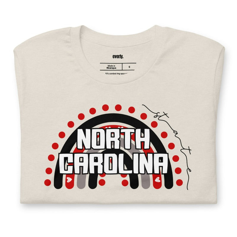 This vibrant NC State design with "North Carolina" text, rainbow arch, and red dots on a beige shirt.