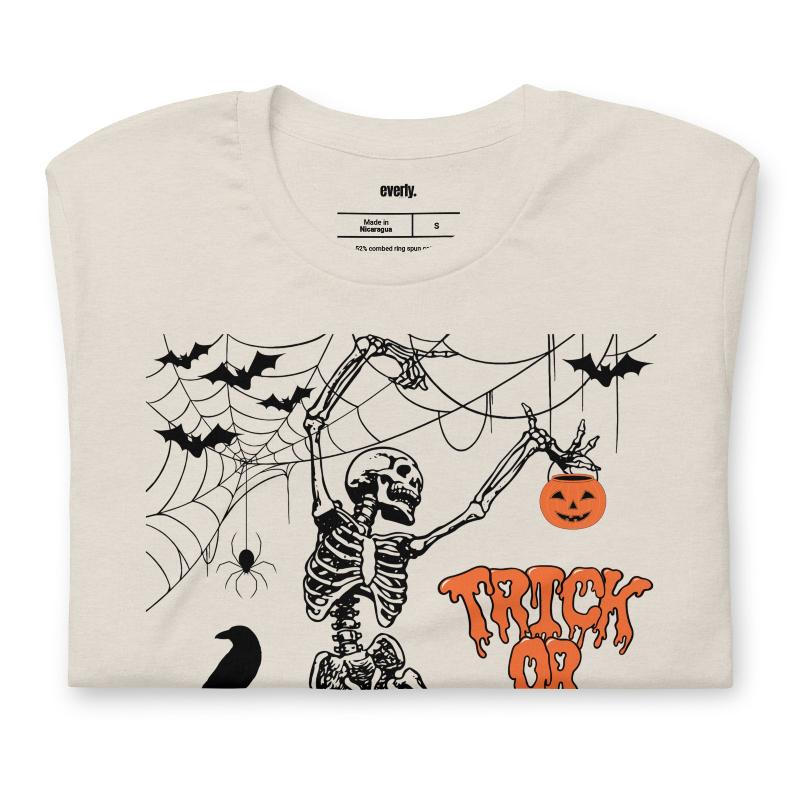 Beige t-shirt with a fun Halloween design featuring a skeleton, pumpkins, bats, and "Trick or Treat" text in a festive orange and black theme.