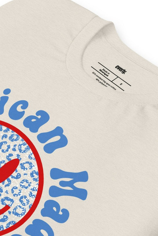 Close-up image of a USA July 4th graphic tee featuring the words 'American Made' surrounded by retro lettering around a bold blue cheetah print retro smiley face on the front. A playful and unique design perfect for celebrating July 4th in style on a heather dust graphic tee.