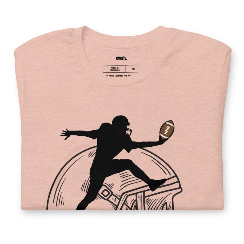 a black silhouette of a football player leaping to catch a football, superimposed on a large, sketched football helmet on a peach shirt. 